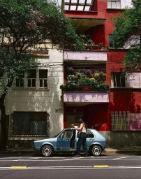 Two people standing by a light blue hatchback car parked on a city street before colourful buildings