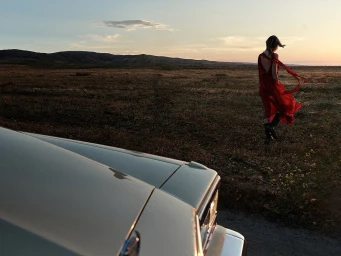 Woman in a red dress standing in a vast grassy field at sunset near the hood of a car, photographed by Anton Watts.