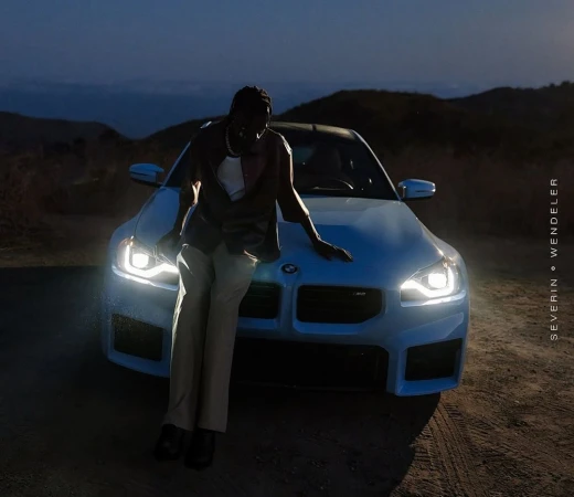 Man leaning against the hood of a blue sports car during dusk with headlights illuminated under a darkening sky.