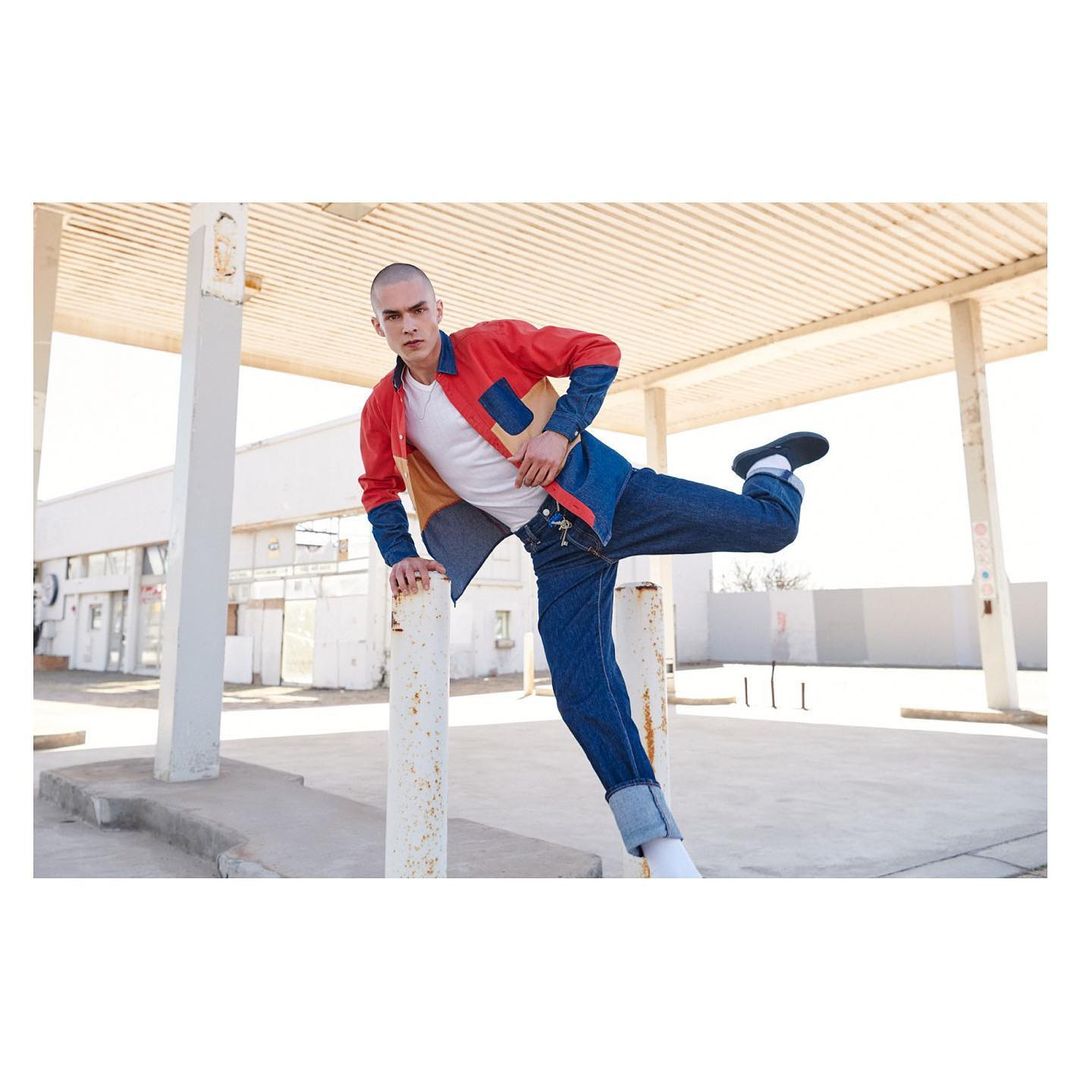An adult male poses at an abandoned gas station wearing a color-blocked jacket and denim jeans, balancing with one leg up on a white pole.