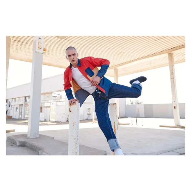 An adult male poses at an abandoned gas station wearing a color-blocked jacket and denim jeans, balancing with one leg up on a white pole.