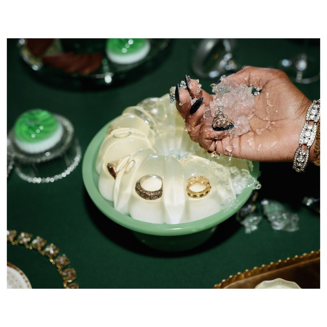 Hand holding rings and crushed ice over jello mold with embedded rings. Jewelry, bracelets, and green tabletop create rich texture.
