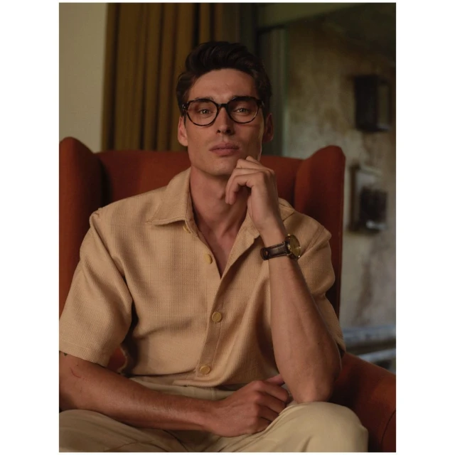Man with glasses wearing a tan shirt, sitting in an orange armchair. He is posing with his hand to his face.