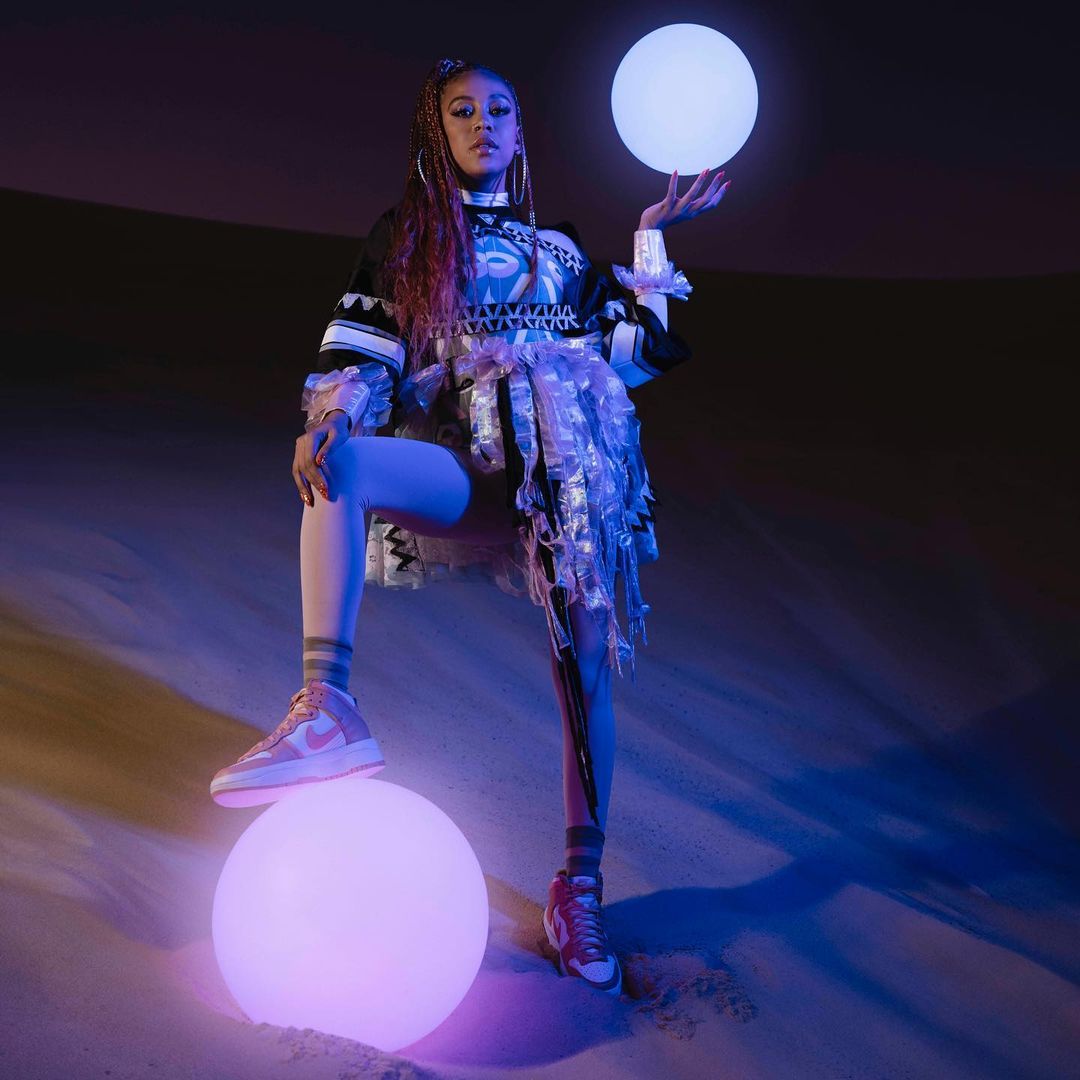 A stylish young woman stands in a desert-like setting, balancing glowing orbs, showcasing a unique fashion ensemble with modern flair.