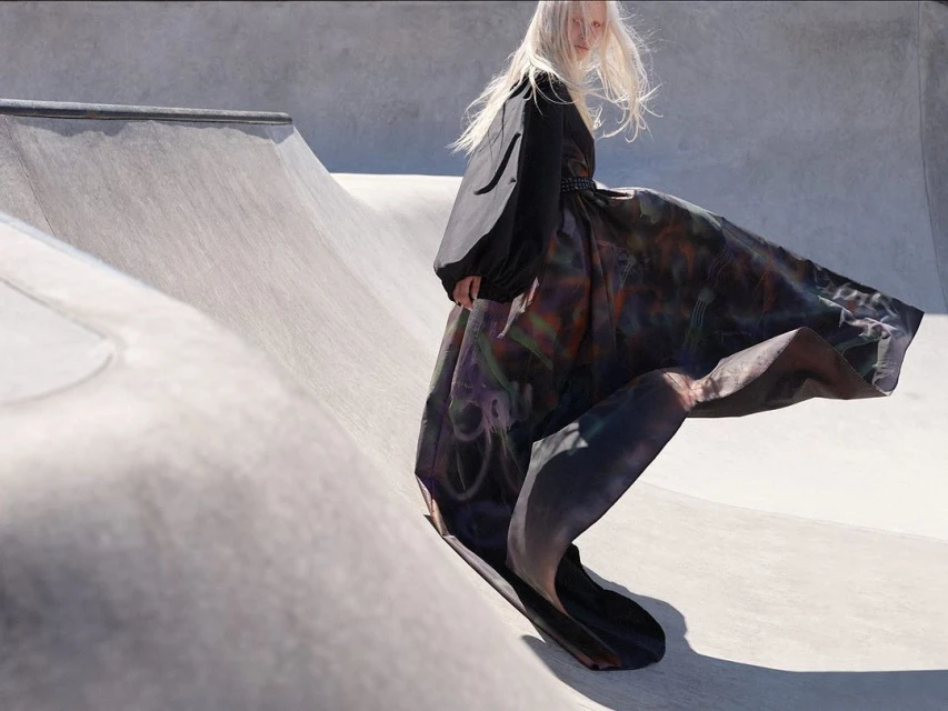 Woman in black top and multicolored dress walking on a concrete skatepark ramp with sunlight, creating a high-fashion editorial image.