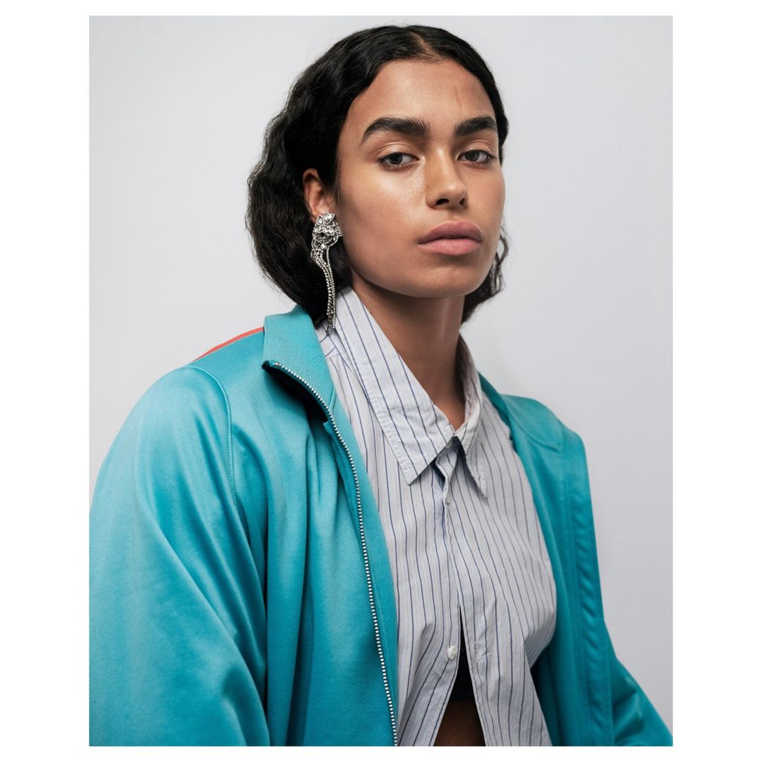 Close-up studio shot of a female fashion model with dark hair wearing a turquoise jacket and a striped shirt, posing in front of a white background.