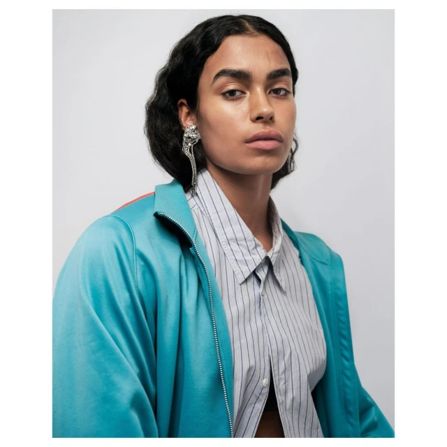 Close-up studio shot of a female fashion model with dark hair wearing a turquoise jacket and a striped shirt, posing in front of a white background.
