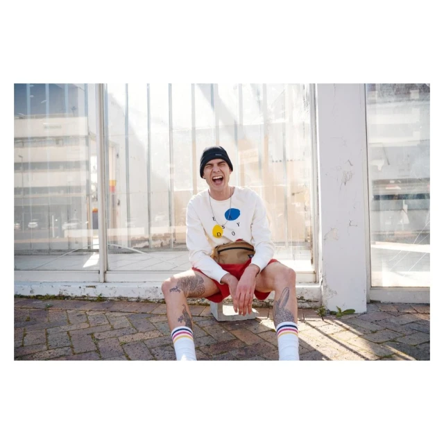 Young adult sitting outdoors on a stool with casual fashion and tattoos, posing with a happy facial expression and street view.