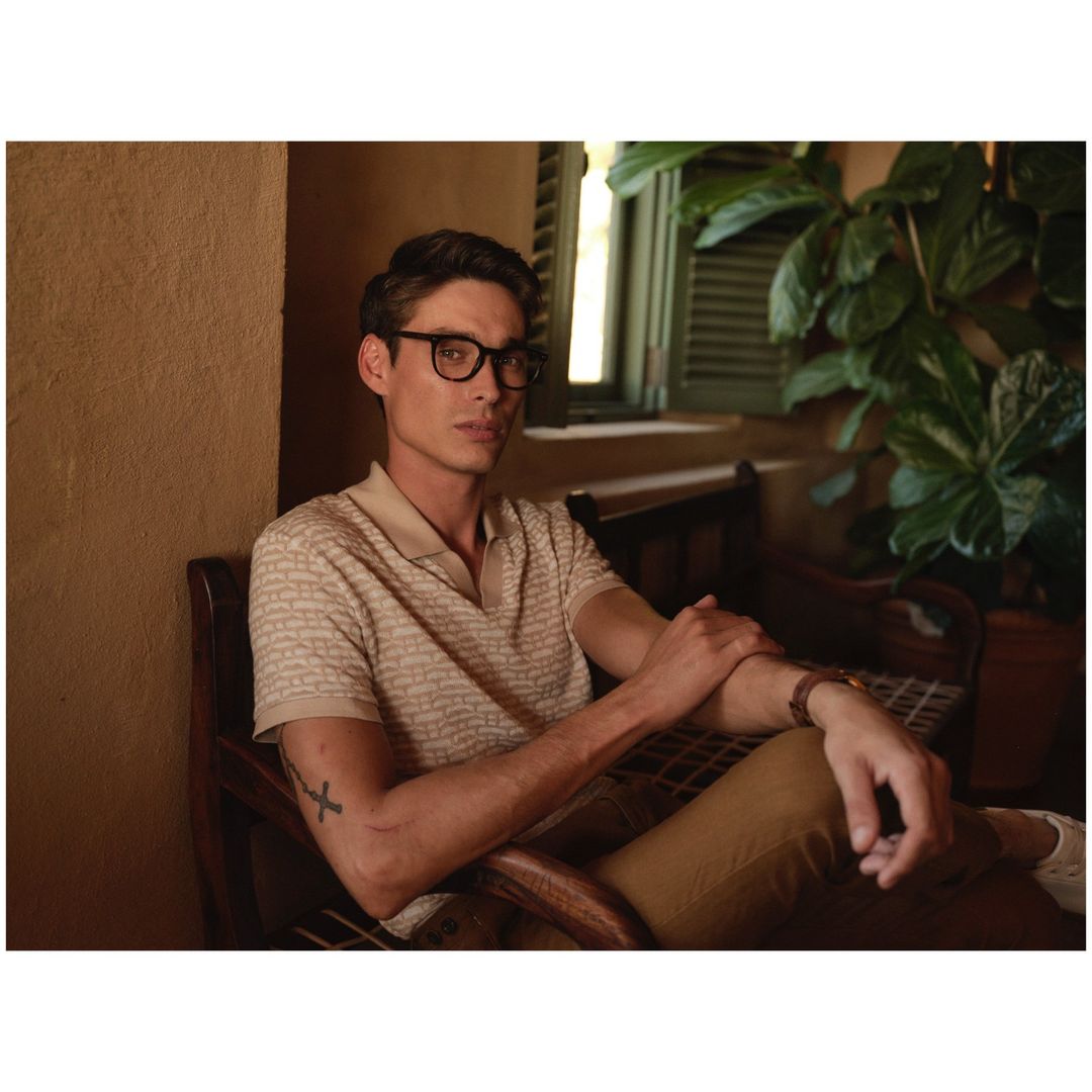 A man sits indoors wearing glasses, a patterned polo shirt, and khaki pants in a warmly lit room, evoking a relaxed lifestyle vibe.