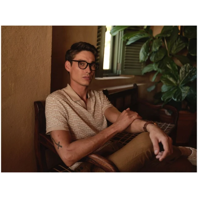 A man sits indoors wearing glasses, a patterned polo shirt, and khaki pants in a warmly lit room, evoking a relaxed lifestyle vibe.