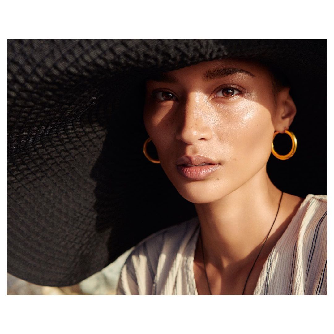 Close-up of a woman with light skin, wearing a large straw hat and gold hoop earrings. The lighting is soft and diffused.