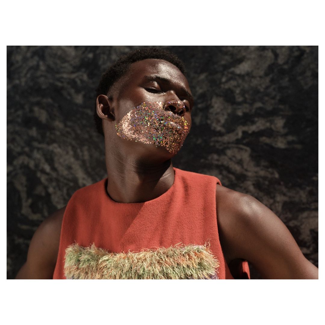 Portrait of Black male model with glitter makeup in rust-colored top against dark patterned backdrop. Fashion and beauty concept.