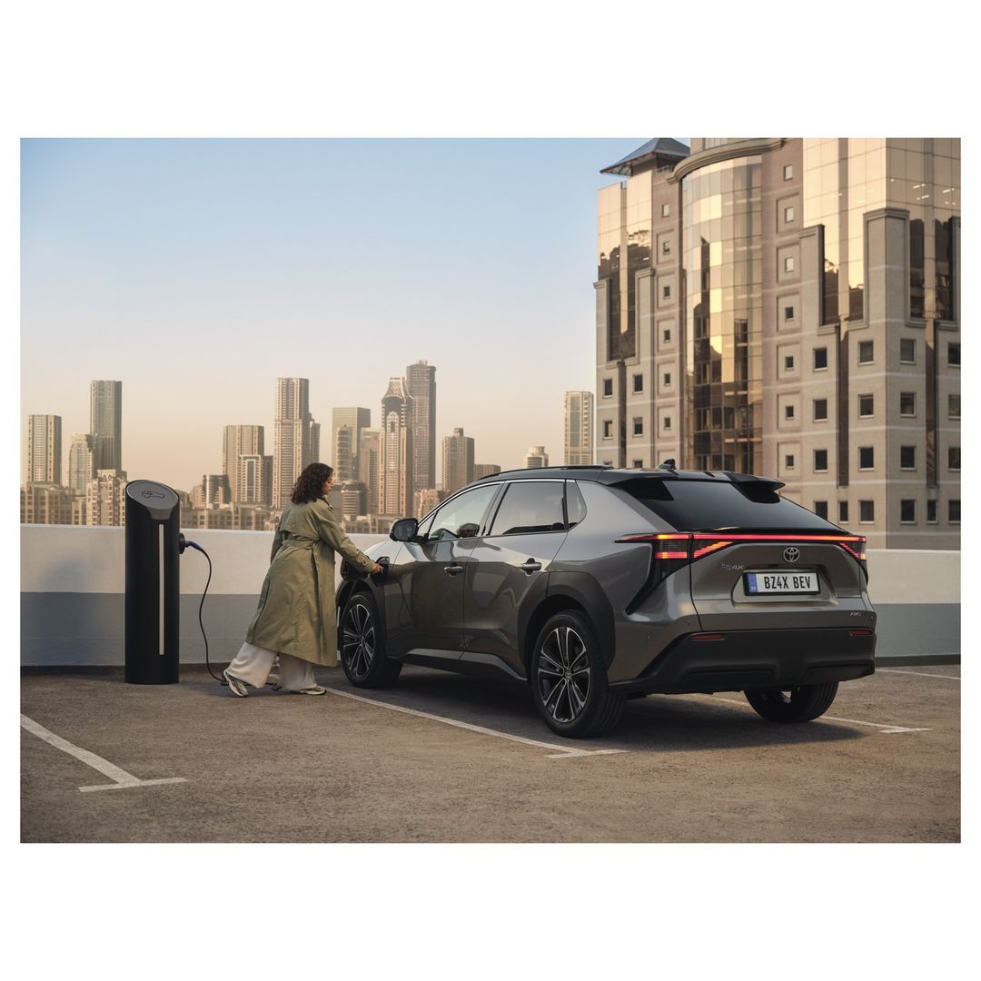 Silver Toyota bZ4X REV parked at a charging station, with a woman plugging in the car. Cityscape and modern architecture in background.