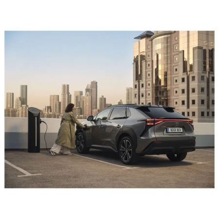 Silver Toyota bZ4X REV parked at a charging station, with a woman plugging in the car. Cityscape and modern architecture in background.
