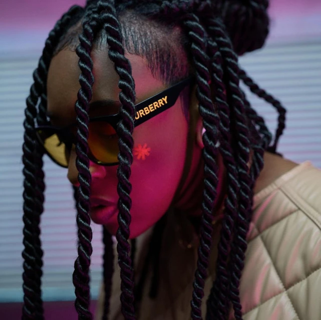 Close up of an adult female with dreadlocks, sunglasses, and a quilted jacket, posing in an indoor environment with pink lighting.