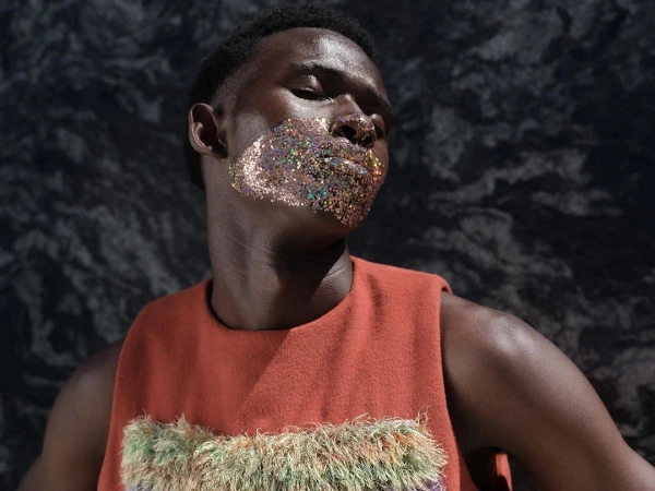 Portrait of a Black man wearing a rust-orange sleeveless top with metallic glitter makeup across his lower face, photographed by Antonia Steyn.