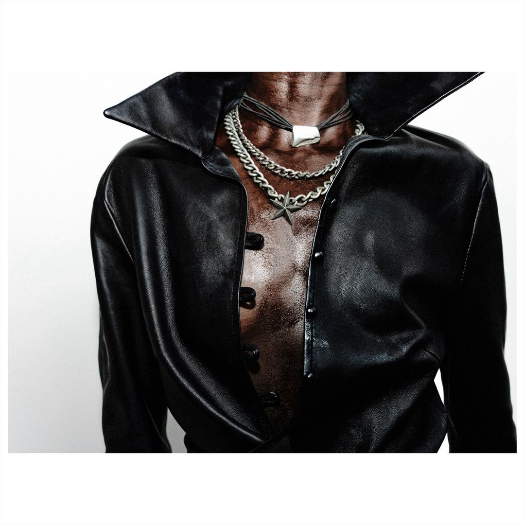 Close-up studio shot of a torso wearing an open black leather jacket, several layered necklaces, and an unbuttoned black collared shirt.