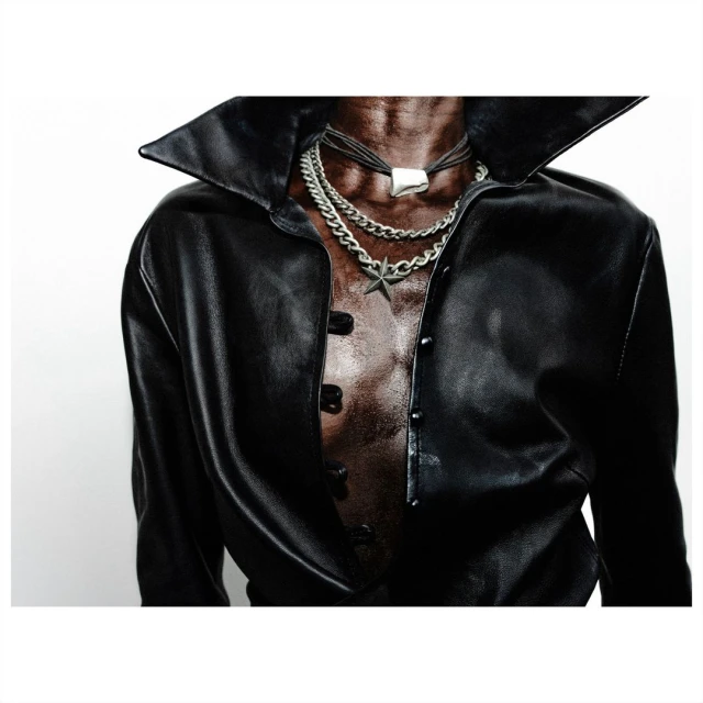 Close-up studio shot of a torso wearing an open black leather jacket, several layered necklaces, and an unbuttoned black collared shirt.