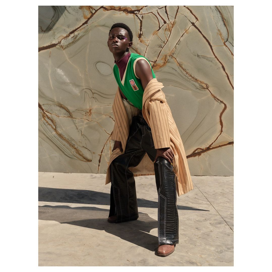 An African male model poses outdoors wearing a green sweater vest, dark pants, a tan coat, and brown shoes against a marble background.