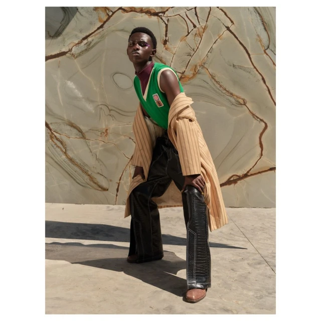 An African male model poses outdoors wearing a green sweater vest, dark pants, a tan coat, and brown shoes against a marble background.