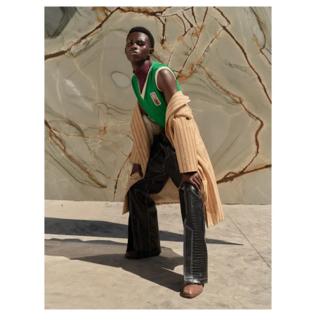 An African male model poses outdoors wearing a green sweater vest, dark pants, a tan coat, and brown shoes against a marble background.