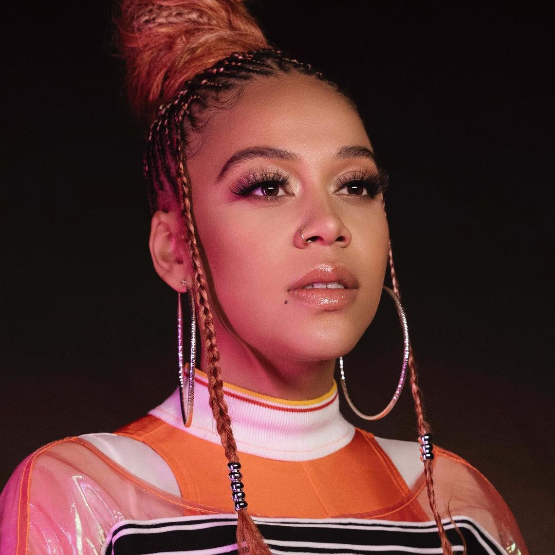 Portrait of a woman with braided hair and hoop earrings wearing an orange top, photographed by Antonia Steyn.