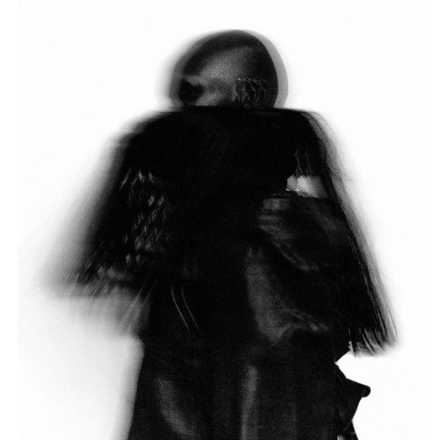 Monochromatic shot of a bald person in dark clothing, obscured by motion blur against a stark white backdrop with visible texture.