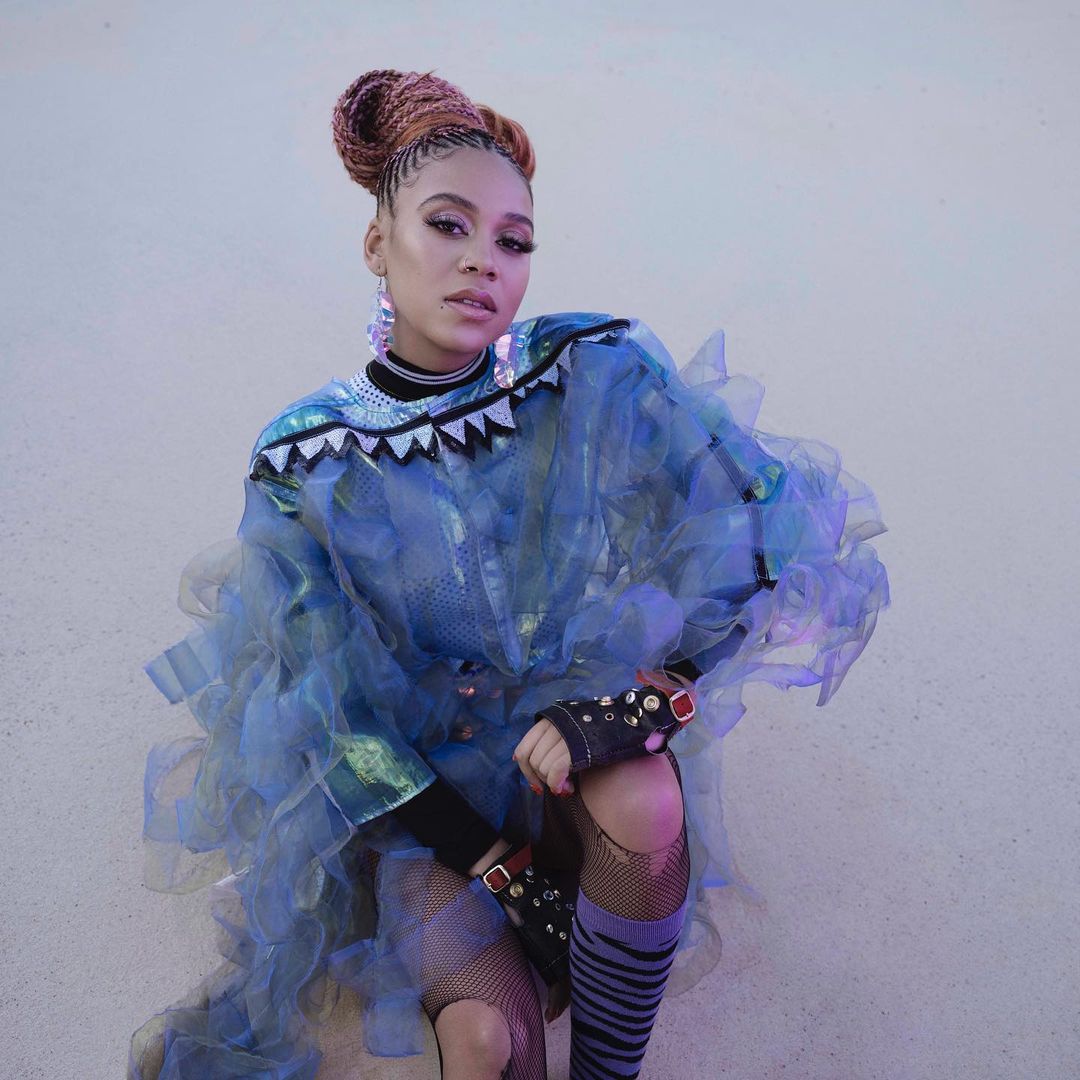 Portrait of a woman with braided hair wearing a blue tulle dress with decorative elements and fishnet stockings in a studio setting.