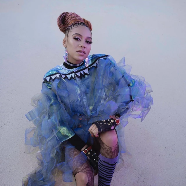 Portrait of a woman with braided hair wearing a blue tulle dress with decorative elements and fishnet stockings in a studio setting.