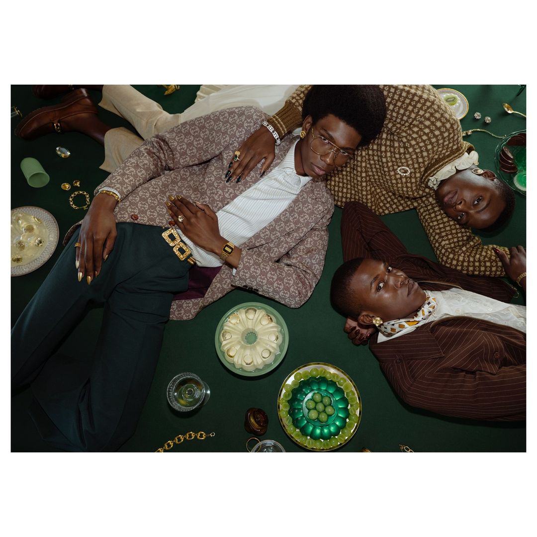 Three Black male models laying on a green surface, wearing blazers, rings, and gold jewelry, with plates of jello and drinks.