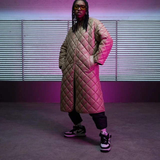 A person with dark skin and dark hair in a tan quilted coat, dark pants, and black and white sneakers stands in a pink and white room.