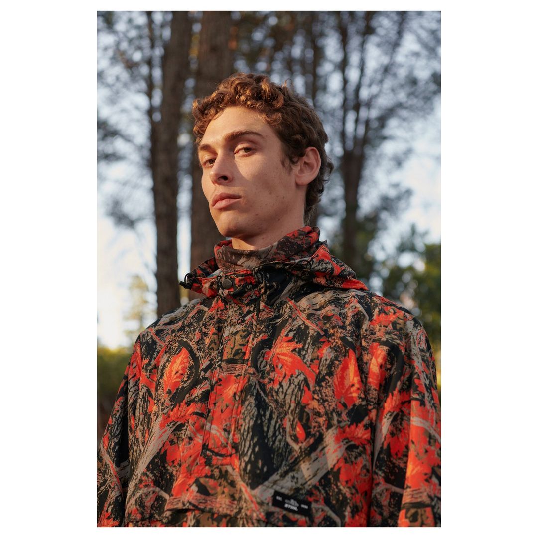 Male model in orange and black camouflage hooded jacket poses in an outdoor woodland setting with trees in the background.