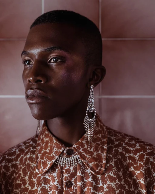Close-up portrait of a black adult male wearing a patterned brown shirt and chandelier earrings with a tiled rose background.