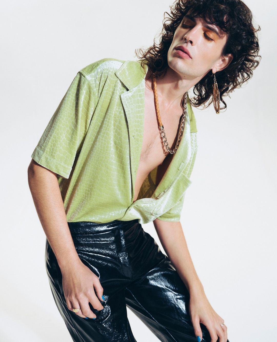 A person poses in a green shirt and black pants in a studio. The individual has dark hair and is accessorized with jewelry.