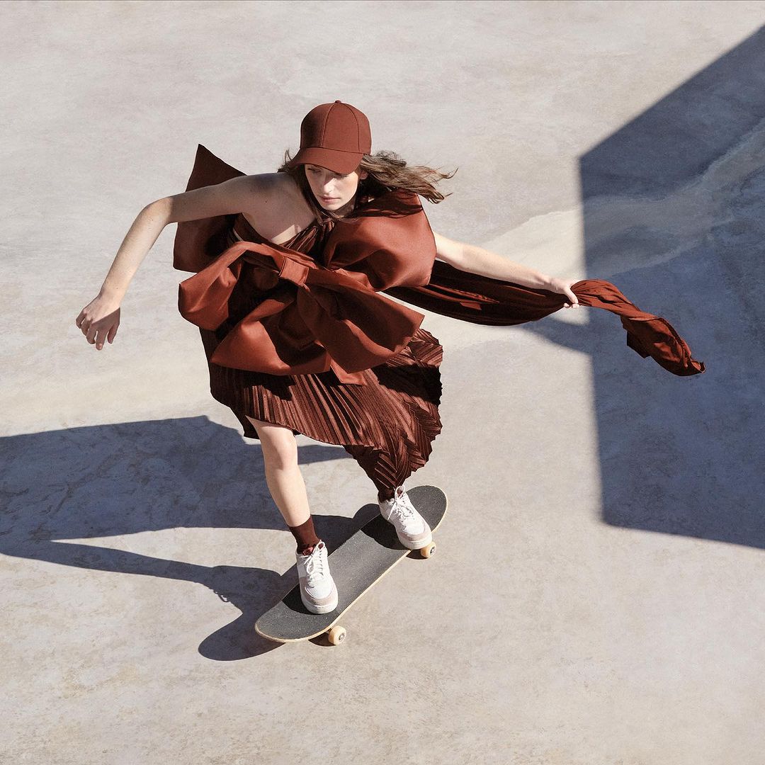 Woman in brown dress and cap skateboarding on concrete, shot from above. Dress has a large decorative bow. Fashion and sport concept.