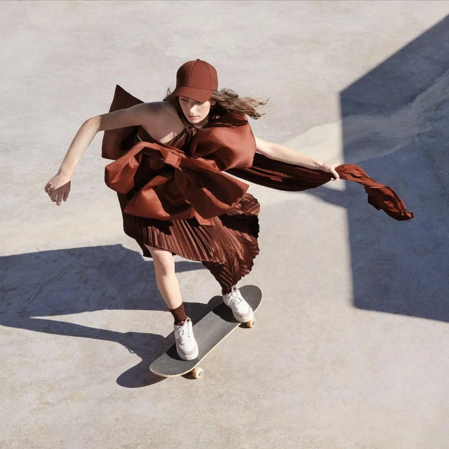 Woman in brown dress and cap skateboarding on concrete, shot from above. Dress has a large decorative bow. Fashion and sport concept.