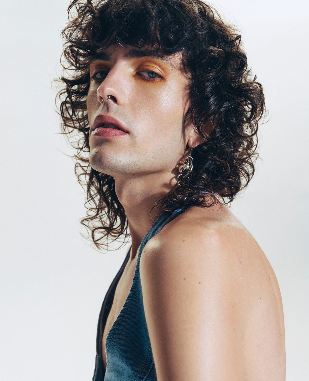 Portrait of an adult male with curly brown hair, orange eyeshadow, nose ring, and silver earrings, wearing a blue top in a photo studio.