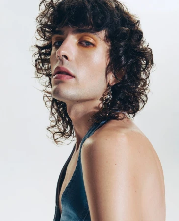 Portrait of an adult male with curly brown hair, orange eyeshadow, nose ring, and silver earrings, wearing a blue top in a photo studio.