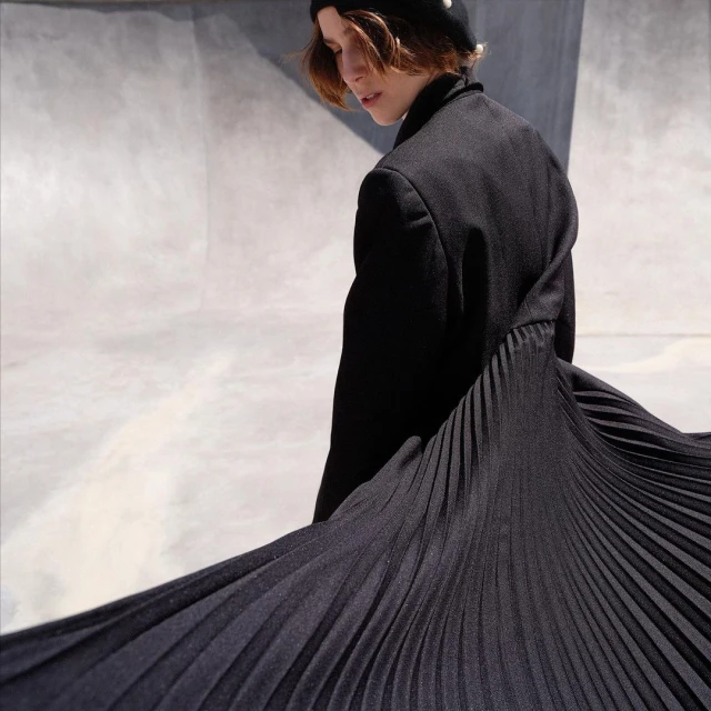 A young woman poses in a black coat with a pleated skirt against a grey wall, looking away from the camera in a fashion setting.