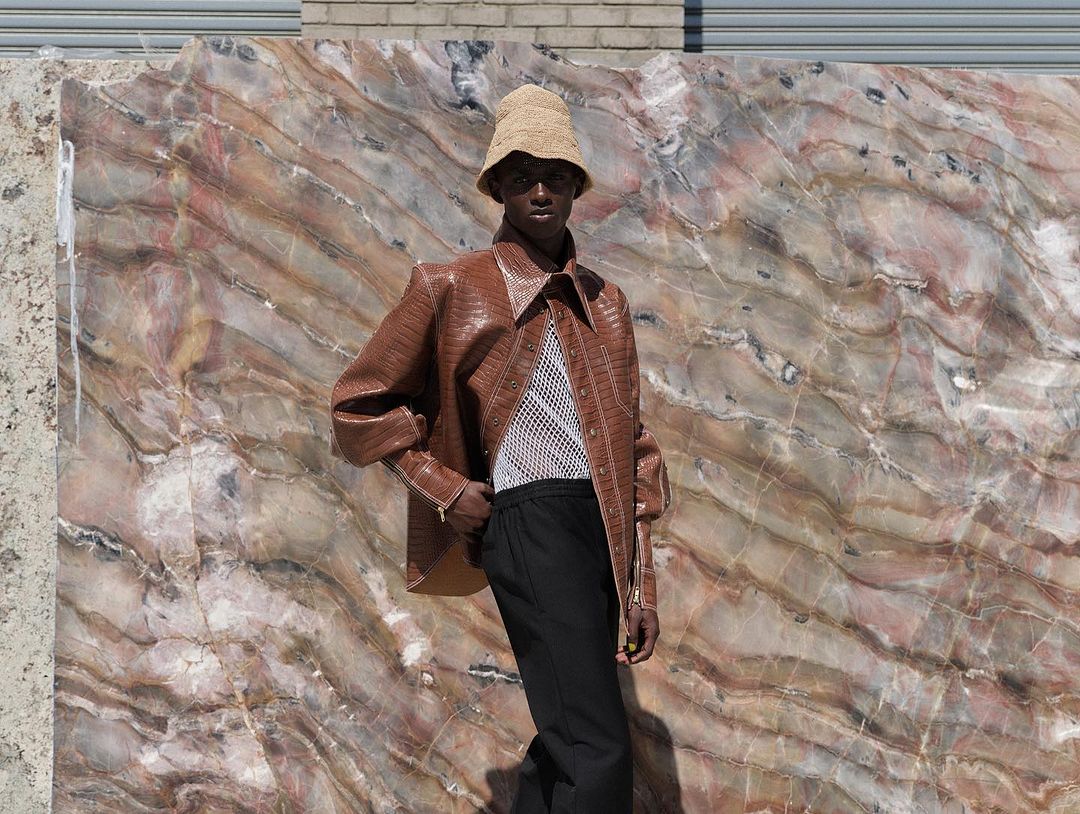 An androgynous fashion model poses in a brown reptile-pattern jacket, black pants, and woven hat against a stone slab background.