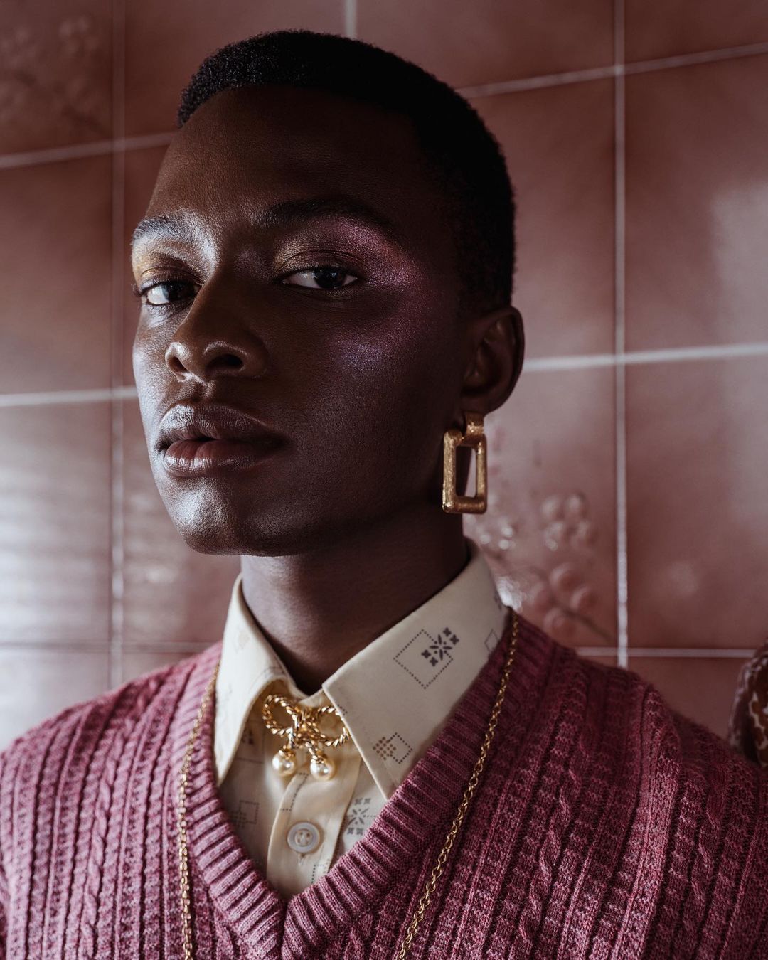 Portrait of a Black man wearing a pink knitted vest and gold earrings against pink tiles by Antonia Steyn
