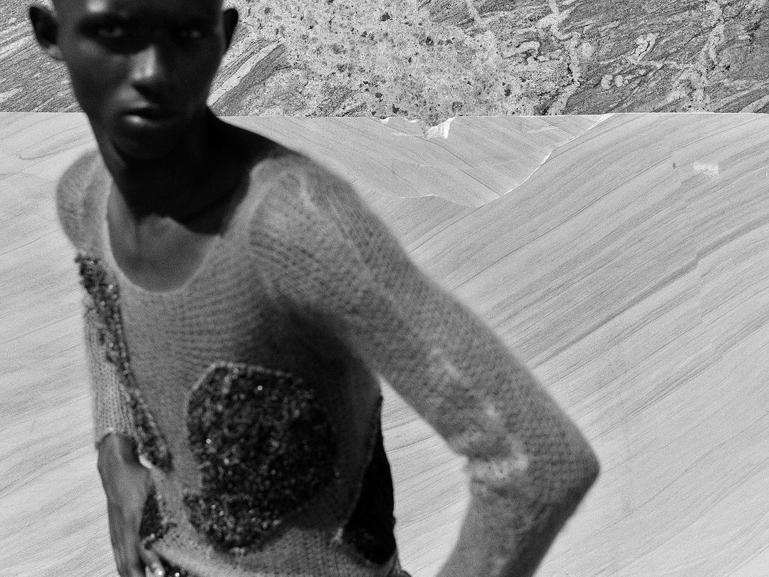 Monochrome portrait of a young Black man in a textured sweater with embellishments, set against a split abstract landscape background.