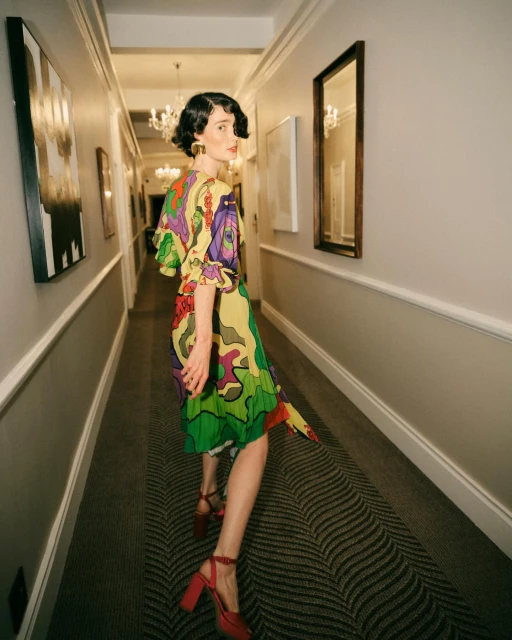 A woman in a yellow, green, and purple printed dress poses in a hallway with art, mirrors, and a chandelier, showcasing a fashion style.