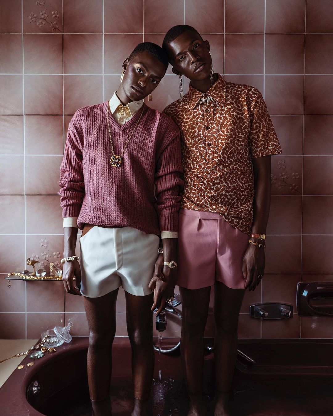 Two adult men pose in a tiled bathroom, adorned with jewelry, wearing fashionable clothing and jewelry while standing in the bathtub.