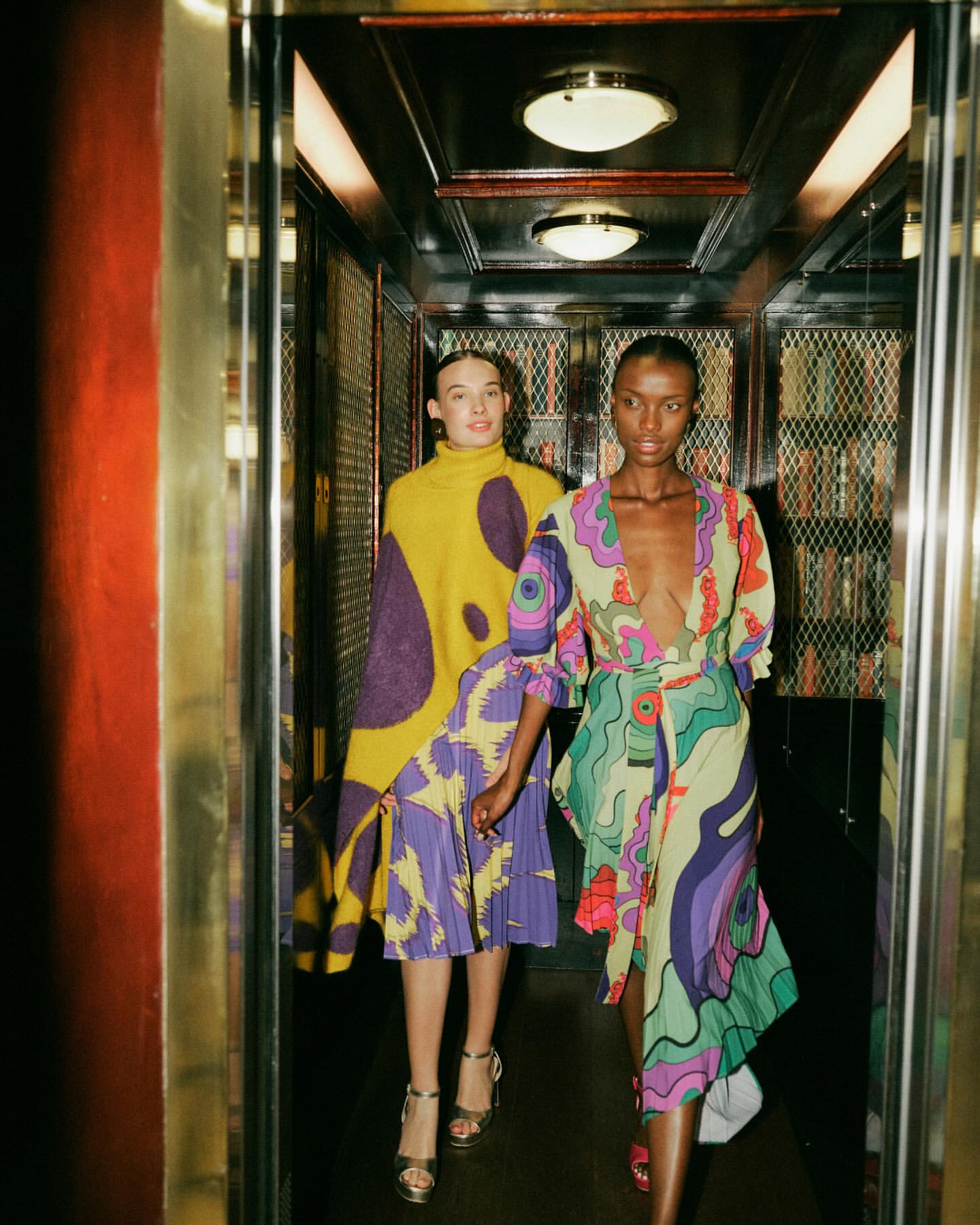 Two women in patterned dresses posing in an elevator with gold trim and mirrored walls, showcasing a vibrant fashion editorial.