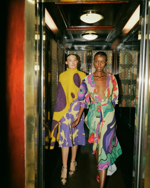 Two women in patterned dresses posing in an elevator with gold trim and mirrored walls, showcasing a vibrant fashion editorial.