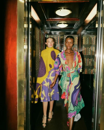 Two women in patterned dresses posing in an elevator with gold trim and mirrored walls, showcasing a vibrant fashion editorial.