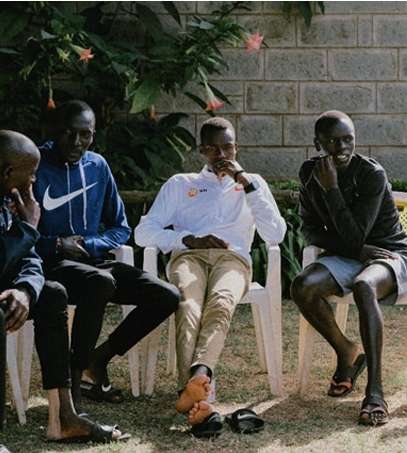 Four men are seated outside in casual clothing, engaged in conversation, with a brick wall and greenery visible in the background.
