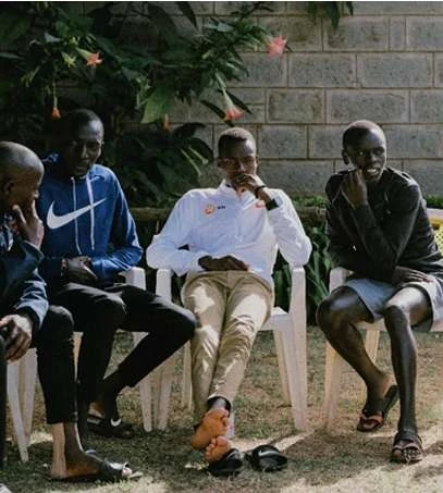 Four men are seated outside in casual clothing, engaged in conversation, with a brick wall and greenery visible in the background.