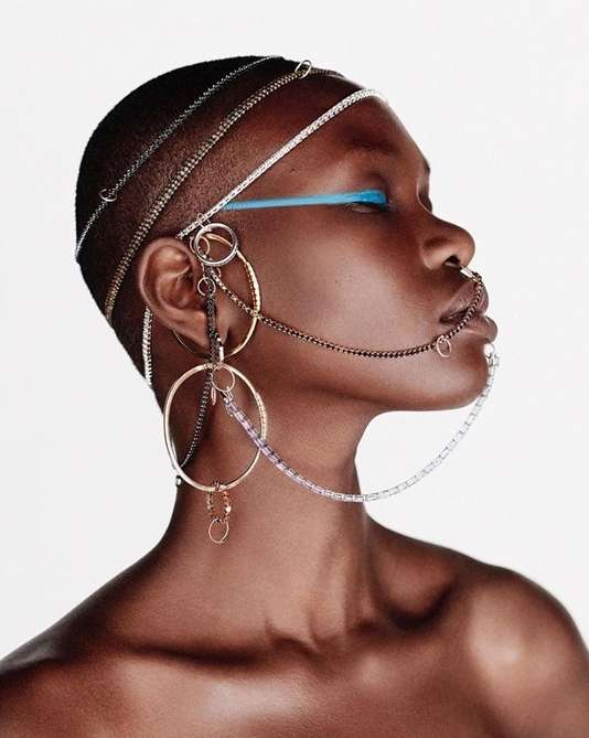 Profile portrait of a fashionable Black woman with short hair wearing distinctive gold and silver jewelry against a bright white backdrop.